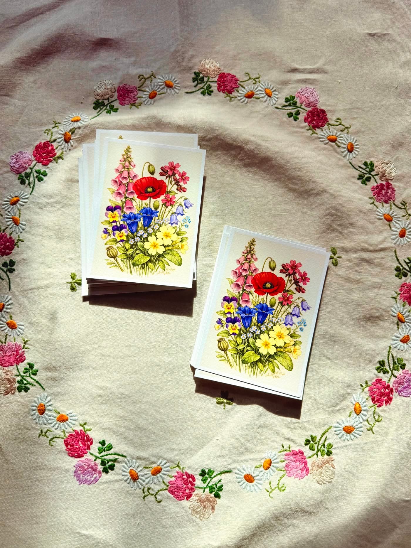 A botanical postcard with a wildflower bouquet — foxglove, poppy, gentian, primrose, pansies — on cotton rag paper resting on embroidered linen.