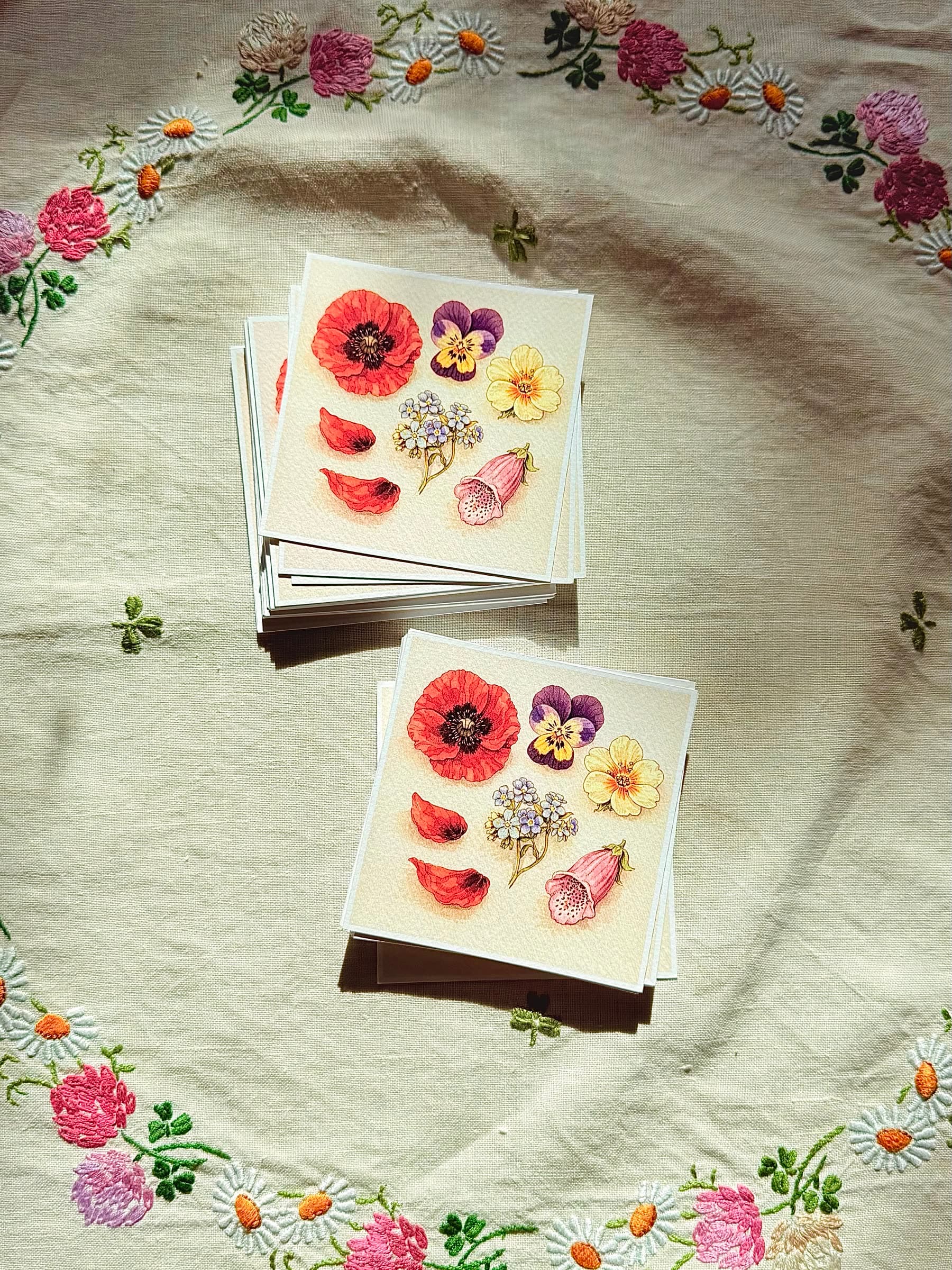 A square art print of a wildflower bouquet — poppy, pansy, primrose, forget-me-nots, foxglove, fallen petals — on cotton rag paper resting on embroidered linen.