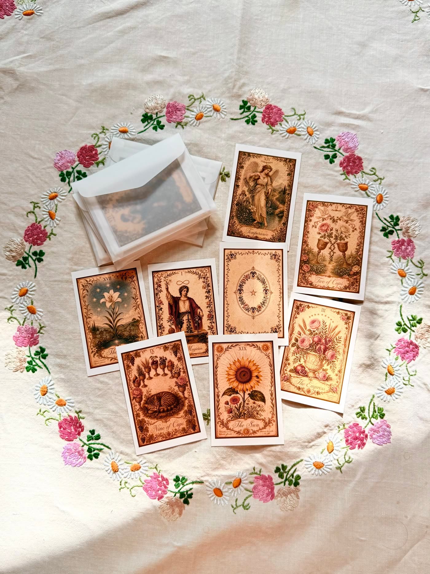 Seven cards from The Garden of Good Omens spread on embroidered linen — Strength, The Sun, The Star, Six of Cups, Three of Cups, Nine of Cups, and a card back with botanical border.